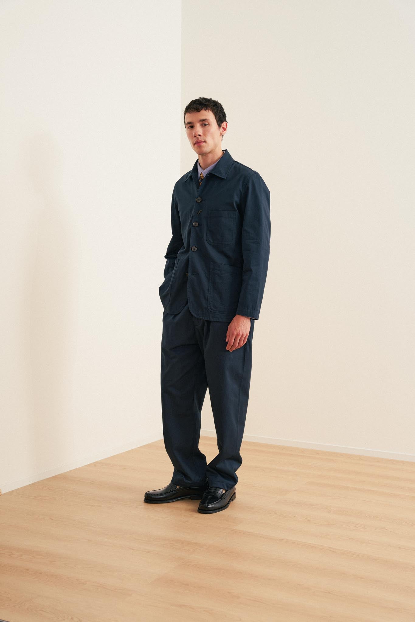 Universal Works Bakers Jacket en sarga de algodón azul marino, llevada con camisa Oxford y pantalón de sarga